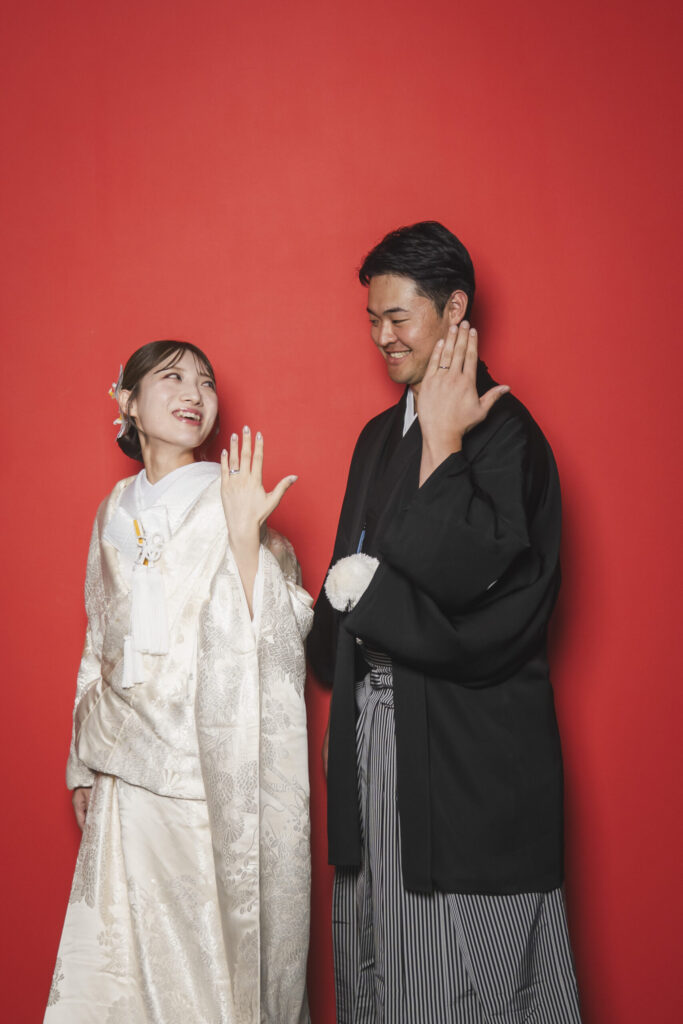赤い壁の前の笑顔で指輪を見せる和装のお二人