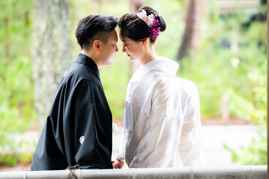 緑を背景に顔を寄せ合う白無垢の新婦と紋付袴の新郎