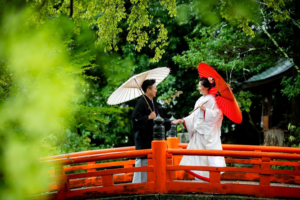 今宮神社の朱色の橋のそばで番傘を持ち見つめ合う新郎新婦
