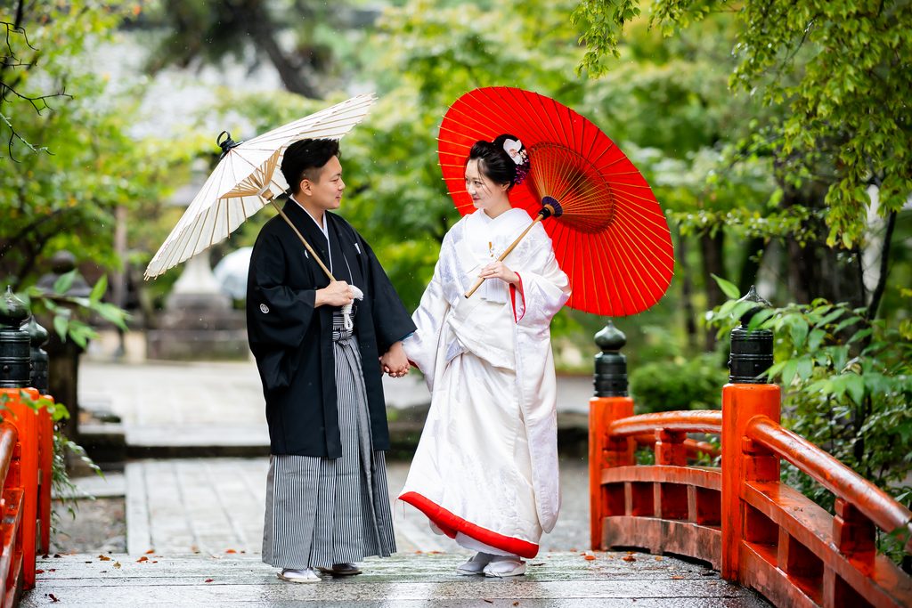今宮神社の朱色の橋のそばで番傘を持ち見つめ合う新郎新婦